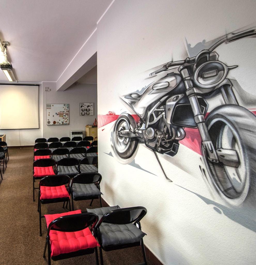 Ein großes, von Hand gemaltes Motorrad an der Wand eines Theorieraums, davor rote und graue Stuhlreihen.